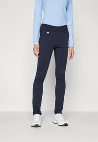 Marineblaue Stretch-Hosen mit glatter Textur, figurbetontem Design, zwei Vordertaschen und einem eleganten Bund, der durch ein rundes Metallelement akzentuiert wird.