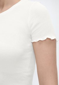 Κοντινό πλάνο ενός ατόμου που φοράει λευκό ριμπ κράμα κοντομάνικο μπλουζάκι με βολάν στις άκρες των μανικιών, με φόντο ανοιχτόχρωμο.