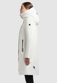 Hvid lang frakke med høj krave og snøre-hætte. Har sorte lynlåse, sidelommer og et logo-patch nær sømmen. Glat tekstur.