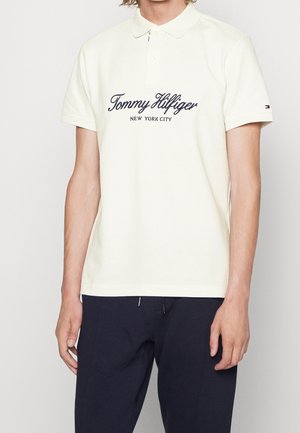 Mężczyzna w białej koszulce polo Tommy Hilfiger z granatowym napisem i czarnych spodniach, stojący na jednolitym tle.