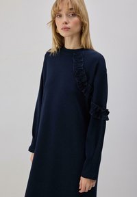 Robe bleu marine avec un col montant, des manches longues et des accents volantés le long du côté gauche et de la manche. Texture de tissu doux.