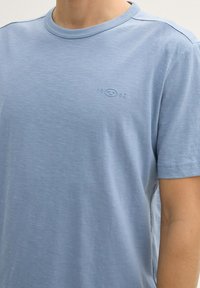 Tricou bleu deschis cu guler rotund, fabricat dintr-un material moale; prezintă un logo subtil embossat cu "1962" pe pieptul stâng. Mâneci scurte și textură netedă.
