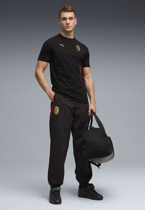 Joven vestido con ropa deportiva negra del AC Milan, sosteniendo una bolsa de viaje negra, sobre un fondo degradado gris.