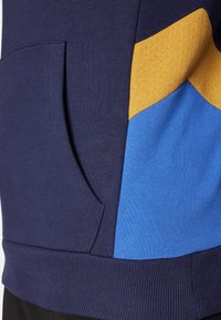 Sweatshirt azul-marinho com um bolso, apresentando acentos triangulares em azul e dourado. O material tem um acabamento texturizado com punhos canelados.