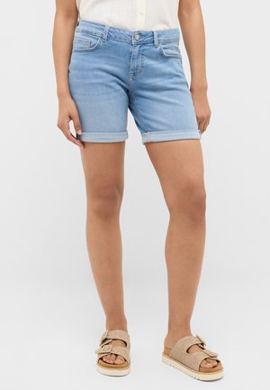 Woman wearing light blue rolled-up denim shorts and beige buckle sandals, standing against a plain white background.