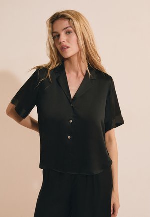 Femme blonde portant une chemise noire ample à manches courtes boutonnée et un pantalon noir, debout devant un fond beige uni.