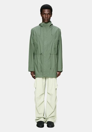 Mężczyzna ubrany w zieloną kurtkę parka z kapturem i sznurkami oraz szerokie, kremowe spodnie typu cargo, stojący na jasnym, jednolitym tle.