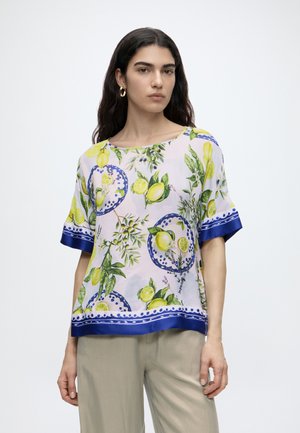 Mujer con blusa blanca con estampado de limones amarillos y hojas verdes y ribete azul, de pie frente a un fondo claro y liso.