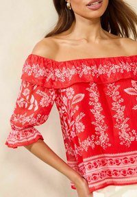 Rød off-shoulder bluse med blomsterprint, lagdelt flæse-detalje og udvidede ærmer, med detaljeret hvidt print og tekstureret stof.