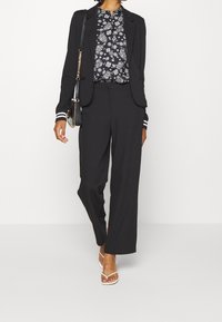 Blazer noir ajusté avec des poignets à rayures blanches contrastantes, associé à un blouse floral noir et blanc et un pantalon large. Sandales blanches à brides.