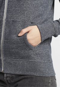 Sudadera con capucha en gris oscuro con una apariencia texturizada, que presenta un cierre frontal con cremallera y bolsillos laterales. Tejido suave y de estilo casual.