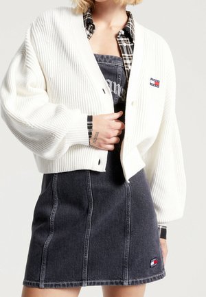 Kobieta nosząca biały prążkowany kardigan na czarnej dżinsowej sukience i koszuli w kratę, z małymi logo Tommy Jeans na kardiganie i spódnicy.