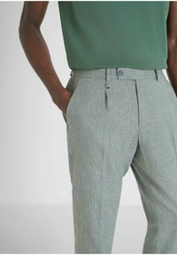 Pantaloni a righe verde chiaro con pieghe, vestibilità sartoriale, tasche laterali e un accento metallico sulla vita, abbinati a una maglietta verde.
