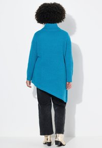 Pull en maille texturée bleu avec un col montant et un ourlet asymétrique. Associé à un pantalon noir et des bottines métalliques.