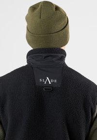Giacca in pile nera con colletto alto, dotata di una toppa in tessuto nera con la scritta "SLADE" e un berretto lavorato a maglia verde oliva.