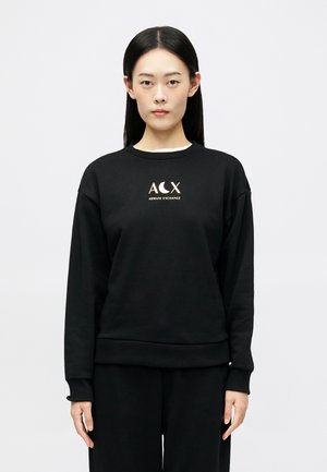 Joven con cabello largo y oscuro, vistiendo un sudadera negra de Armani Exchange con el texto del logo, de pie contra un fondo claro y sencillo.