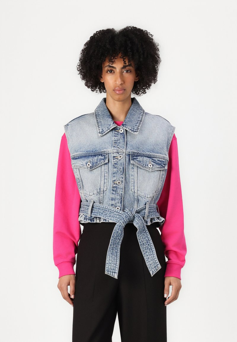 Gilet in denim con vita cintata di colore azzurro chiaro, dotato di due tasche sul petto e di un colletto, indossato sopra una maglietta a maniche lunghe di un vivace colore rosa.