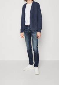 Cárdigan azul marino con una textura suave, cierre de botones, llevado sobre una camiseta blanca, combinado con jeans de mezclilla oscura y zapatillas blancas.