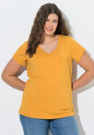 Junge Frau mit langen lockigen Haaren, die ein gelbes V-Ausschnitt T-Shirt und blaue Jeans trägt, steht mit den Händen hinter dem Rücken.