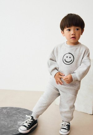 Szara bawełniana bluza z czarną uśmiechniętą buźką, w połączeniu z pasującymi spodniami dresowymi. Dziecko ma na sobie czarne sneakersy z białymi akcentami.
