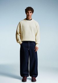 Hellbeige, gerippter Strickpullover, lockere, dunkle Jeanshose und rote Sneakers. Das Model steht vor einem verlaufenden grau-blauen Hintergrund.