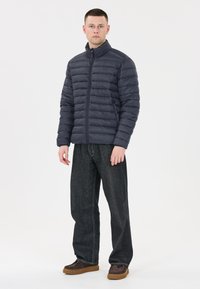Mörkgrå dunjacka med horisontellt quiltad design, hög krage och dragkedjestängning, i kombination med mörka jeans och bruna skor.