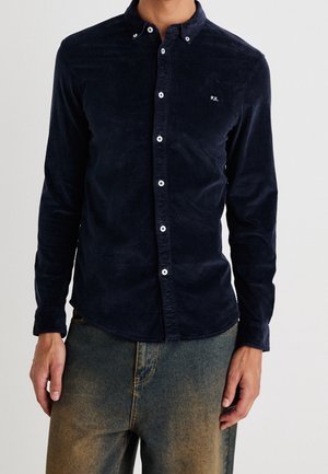 Camisa de pana azul marino con cuello abotonado, botones blancos y pequeño logo bordado en el pecho. Mangas largas, tejido texturizado, diseño ajustado.