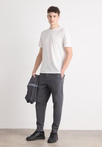 Pantaloni de trening gri cu detalii texturate, hanorac gri asortat cu o dungă albastră și un tricou gri deschis. Modelul stă pe un fundal neutru.