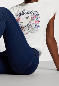 Persona vestida con jeans azul oscuro y una camiseta blanca con una bicicleta floral colorida y el texto "Vacaciones", sentada con una rodilla levantada.