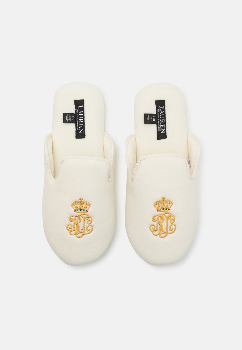 Des pantoufles blanches à enfiler avec une texture douce, arborant un motif de monogramme doré brodé et un design de couronne, étiquetées "LAUREN" sur des étiquettes noires.