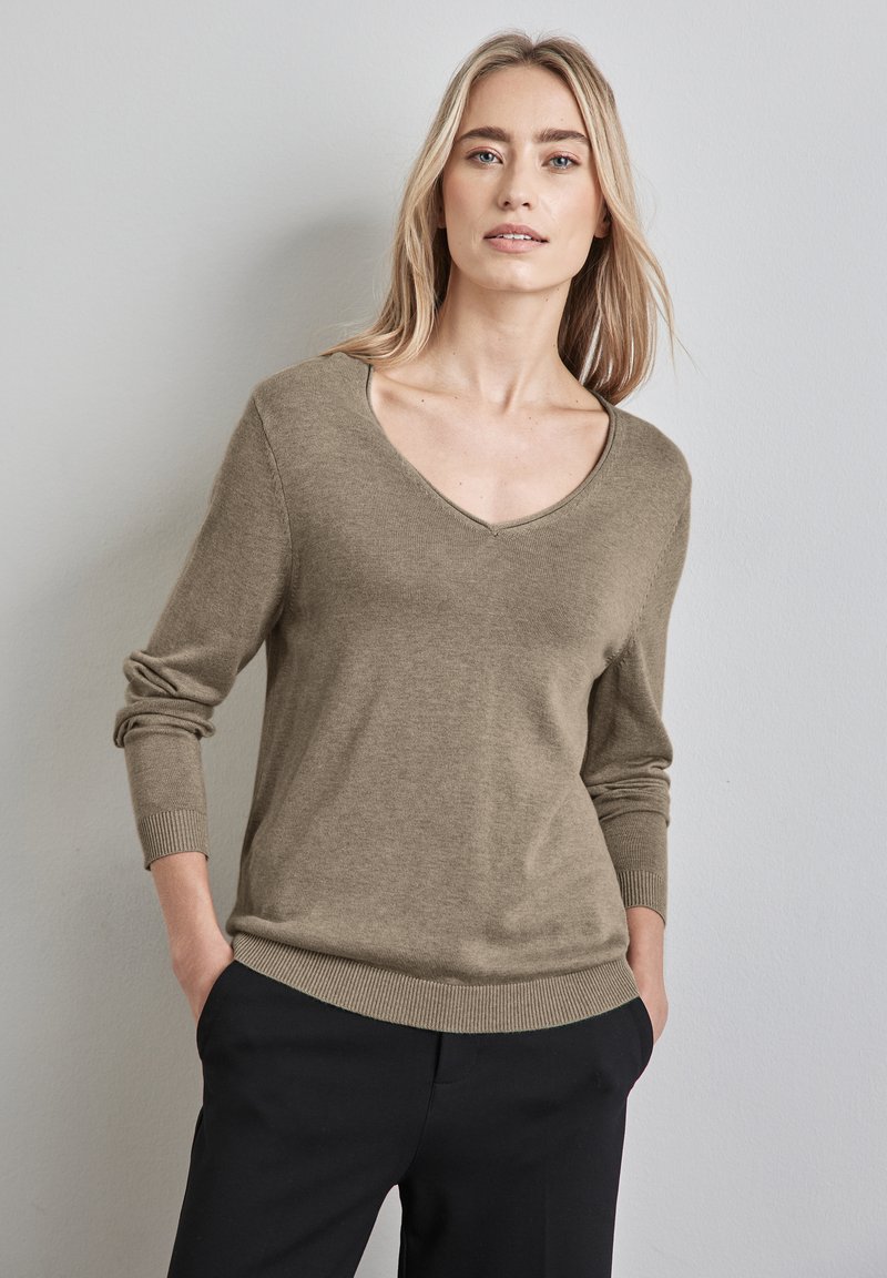 Beige V-ringad tröja i ett mjukt material med långärmar och ribbade muddar, i kombination med svarta byxor. Slät yta, avslappnad design.