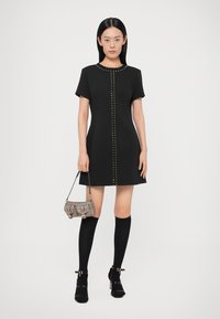 Černé šaty s krátkými rukávy a přímým střihem, zdobené zlatými cvočky ve středu. Kombinovány s černými podkolenkami po kolena a malou stříbrnou kabelkou.