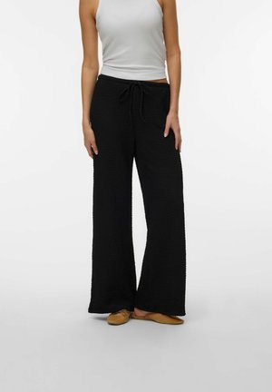 Kvinde iført sorte teksturerede bukser med vide ben og snøre i taljen, hvid ærmeløs top og beige flade sko på ensfarvet baggrund.