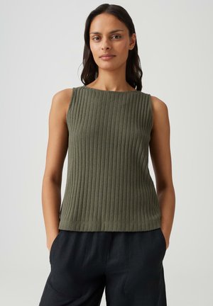 Femme aux longs cheveux foncés portant un haut sans manches côtelé vert olive et un pantalon noir, debout les mains dans les poches devant un fond uni.