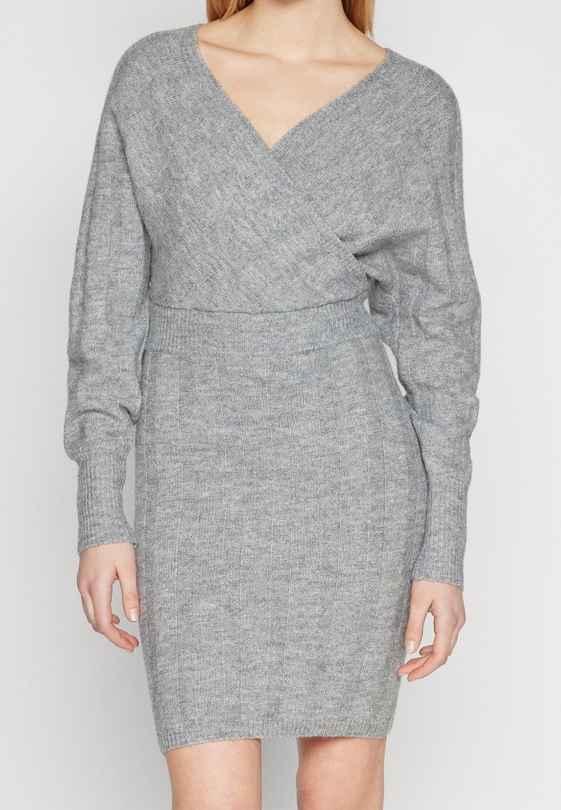 Femme portant une robe en maille gris clair ajustée avec des manches longues et un corsage style portefeuille en V.