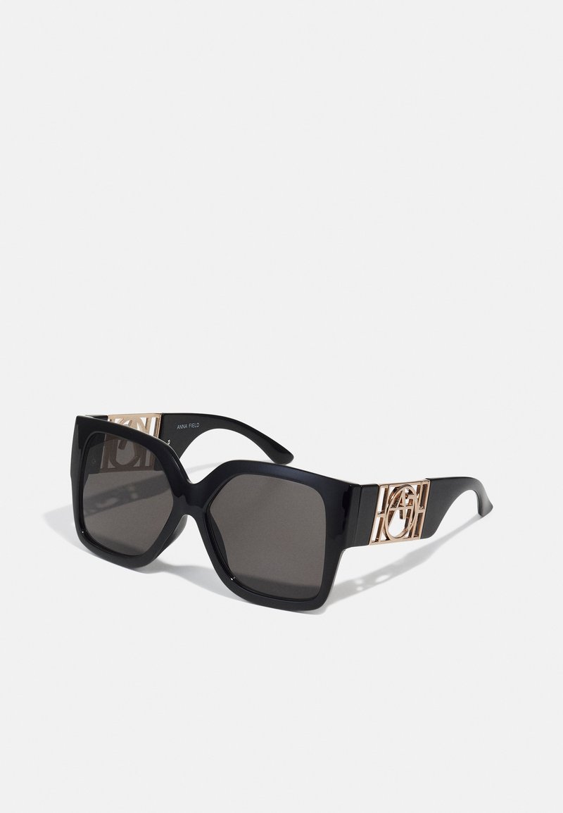 Gafas de sol negras de gran tamaño con lentes oscuros, que presentan un diseño de marco geométrico y acentos en tono dorado en las patillas.