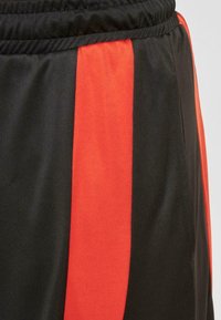Black athletic shorts featuring red side stripes, elastic waistband, smooth texture, with side pockets and stitching details visible.