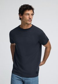 Camiseta de algodón de manga corta en azul marino con cuello redondo y parche de logo sutil. Textura suave y corte relajado. Combinada con jeans de denim claros.