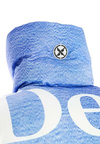 Chaqueta acolchada azul con textura vaquera, cuello alto y grandes letras blancas. Logotipo circular negro con detalles de diseño en la parte trasera.