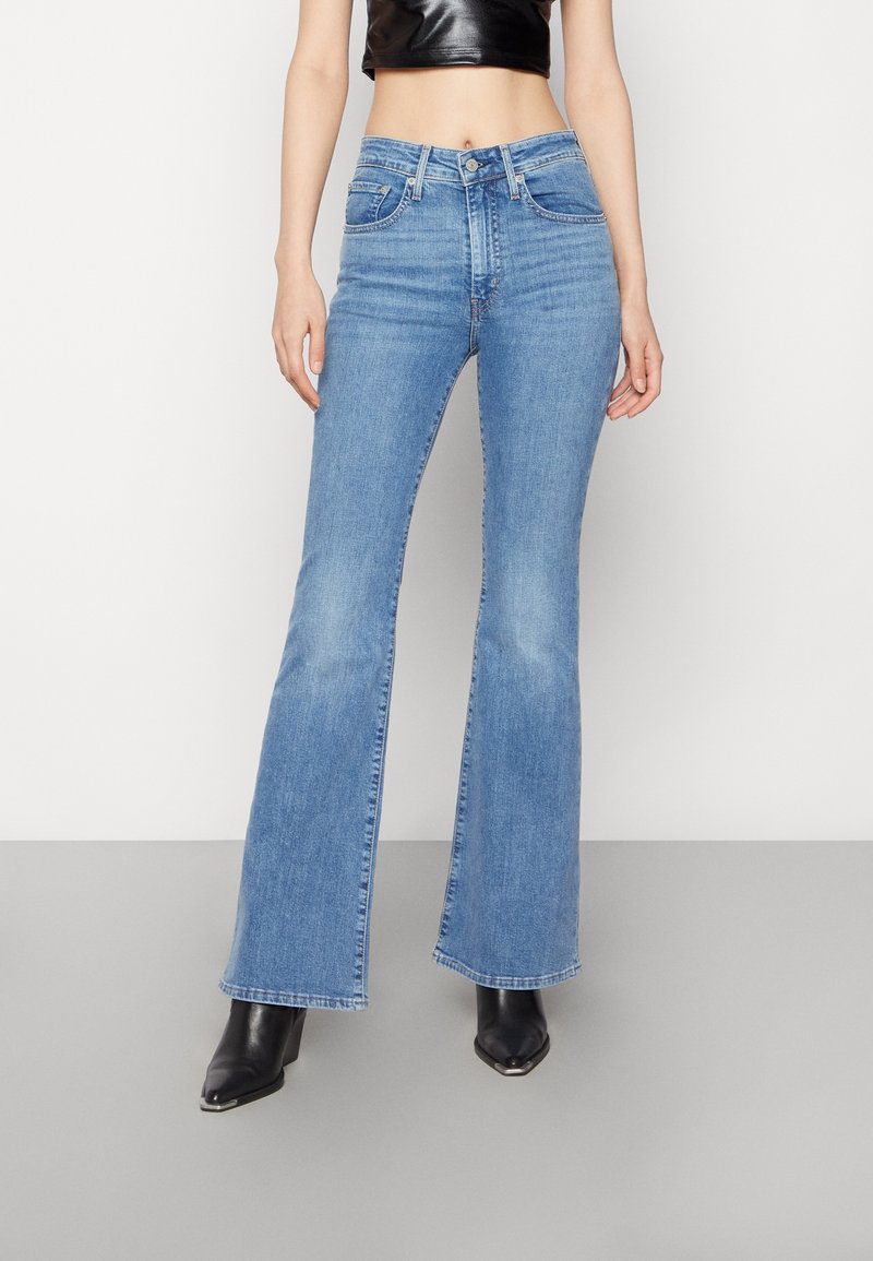 Femme portant un jean évasé taille haute bleu clair et des bottes noires pointues, debout devant un fond clair uni.