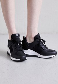 Černé sportovní boty s kombinací texturovaných materiálů, doplněné o bílé detaily, tkaničky a mírný podpatek pro oporu.