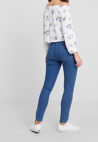 Top blanco sin mangas con un patrón floral azul, combinado con unos jeans ajustados de cintura alta en azul oscuro y zapatillas blancas. Textura suave.