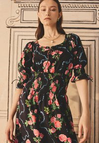 Rochie neagră cu flori, având trandafiri roz și frunze verzi, cu un corp elastic, mâneci bufante și detalii albastre pe modele florale.