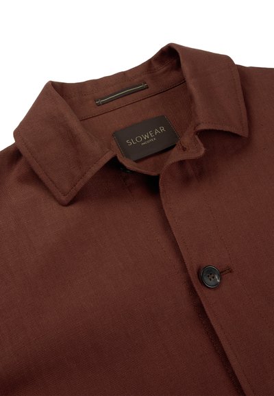 Brązowa lniana koszula z ostrym kołnierzykiem, czarnym guzikiem i czarną metką z napisem "SLOWEAR INCOTEX" wewnątrz kołnierza.