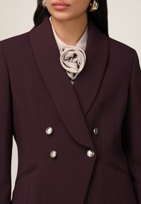 Dvouvrstvý blejzr z tmavě bordó látky, zdobený jemnou béžovou květinovou broží, knoflíky ve stříbrném tónu a střihanými bočními kapsami.