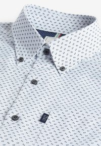 Camisa de cuello blanco con un pequeño patrón de hojas en tono marino, botones oscuros y una etiqueta con un logo gráfico en el bolsillo del pecho.