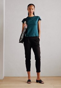 Türkisfarbener Kurzarm-Top mit Rüschendetails, kombiniert mit schwarzen, schmalen Hosen und schwarzen Sandalen, während sie eine große schwarze Tasche hält. Hintergrund mit Holzboden.