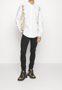 Bílá košile s knoflíky a žlutými grafickými akcenty, černé úzké kalhoty a černé/zlaté tenisky s lesklými detaily.