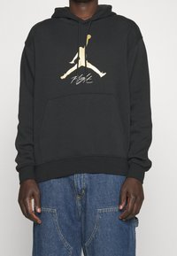 Persona che indossa un hoodie nero con il logo Jumpman dorato e il testo "Flight", abbinato a jeans di denim blu larghi.