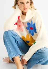 Pullover lavorato a maglia color crema con colletto oversize e applicazioni floreali colorate in rosa, rosso, blu, giallo e verde; abbinato a jeans azzurri chiari.